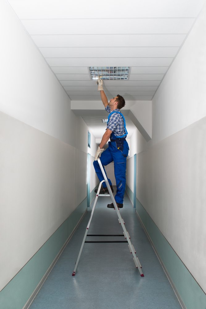 Full length side view of electrician on stepladder installs lighting to the ceiling in office